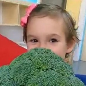 A girl holding up a piece of broccoli covering half of her face.