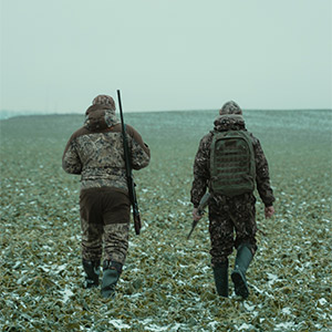 2 hunters carrying rifles walking through a snowy pasture