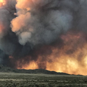 Large plumes of dark smoke are rising from a wildfire in the distance, on the other side of a hill and sagebrush and grass cover the landscape in the foreground.