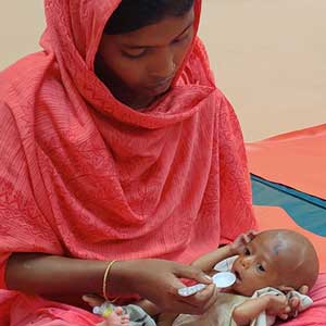 In a hospital, a woman in a bright red shalwar kameez dress and dupatta shawl spoon-feeds an emaciated infant in her lap.