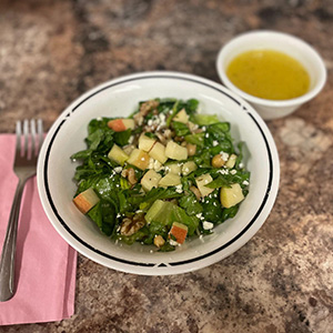 bowl with salad, apple pieces, cheese and small cop with yellow dressing on the side.