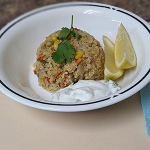 bowl of rice looking food with green garnish on top, lemon slices on the side, white sauce on the side.