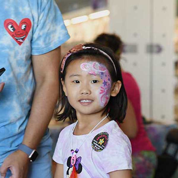 A young girl around age 5 with black hair wearing a headband smiles and shares her pink, white and purple face painting design. 