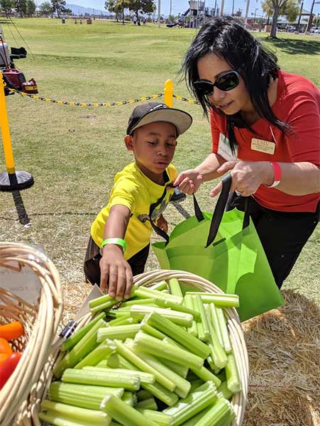 An intern holds a reusable shopping bag open while a child picks healthy produce, including celery, to put into the bag.