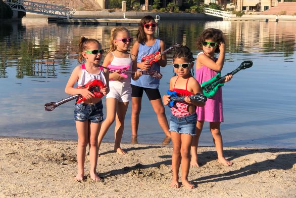 Five girls playing on toy guitars in front of a lake. 