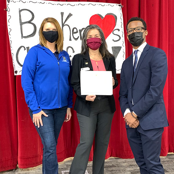 In front of a red stage curtain and homemade Booker Loves Chefs for Kids banner, two Extension staff members stand with a representative from Congressman Horsford's office.