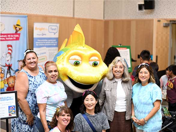 Six women smile while they stand in front of a yellow fish mascot from the PBS show “Splash and Bubbles.”