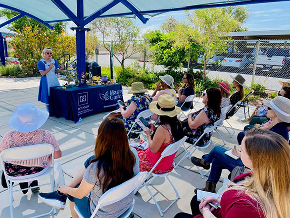 A group of individuals sit in a chairs while listening to a woman in a blue dress and white apron give a presentation outside under a covered pavilion. 