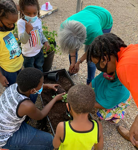 Five youth gathered around Tricia Braxton Perry and a planter box as Tricia explains and demonstrates how to plant in a garden bed. 