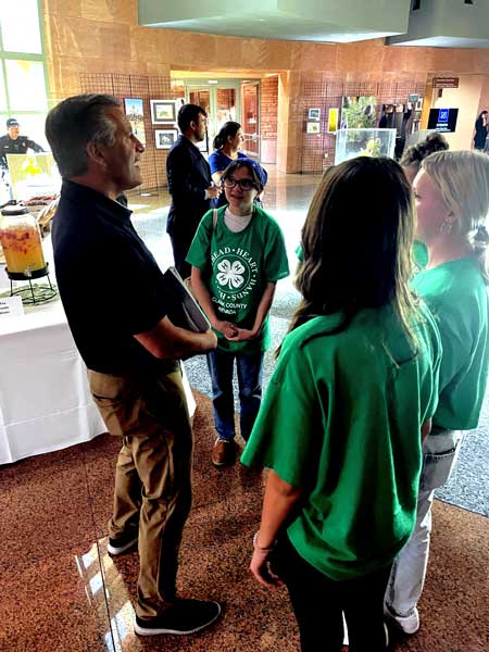 UNR President Brian Sandoval, who is wearing khakis and a navy polo, stands with three youth wearing green 4-H shirts in a semi-circle as they talk at the Extension event.