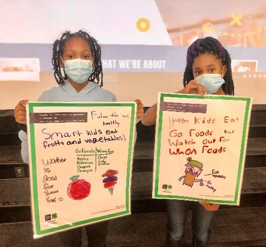 Two elementary aged-girls hold poster boards with their drawings and messages about healthy eating such as go and whoa foods. 