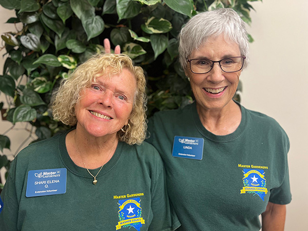 Two people wearing green shirts smiling at camera standing in front of a tree. 