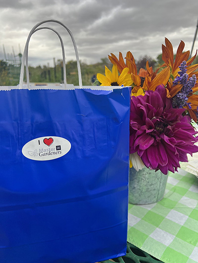 Blue gift bag with Master Gardener logo and purple, yellow and orange flower in the background.