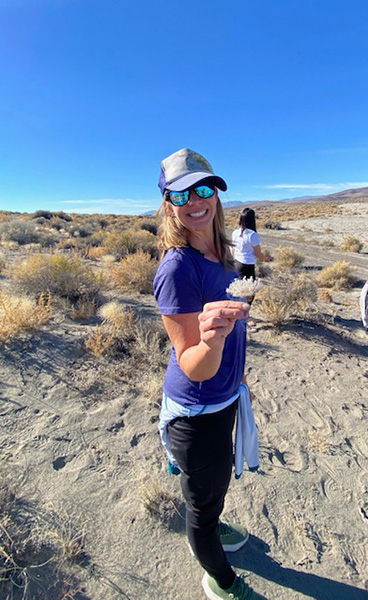 A person wearing a hat, sunglasses and holding a white flower outside standing in front of sage brush and a blue sky in background.