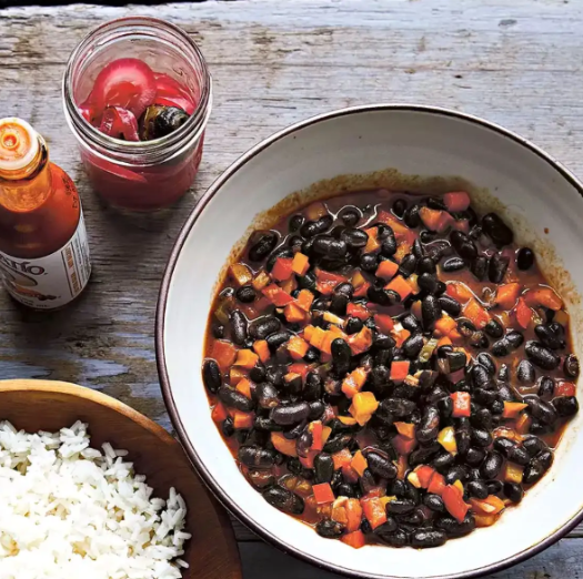 Cuban-Style Orange-Scented Black Beans in a bowl with a side of rice and Tapatio hot sauce. 