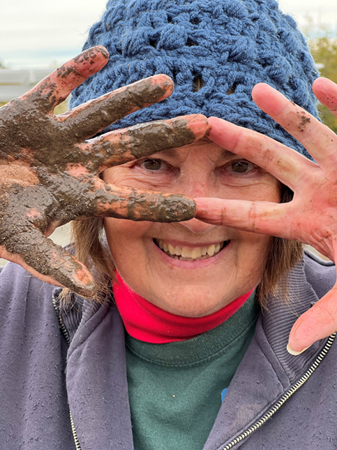 Person wearing a blue hat and smiling with hands with dirt in front of face.