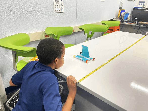 A student measures the distance their air powered vehicle travels down a table.