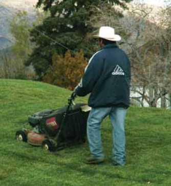 A man with his back facing the viewer mows away down a large lawn, he has a large hat and protective pants and sleeves on.