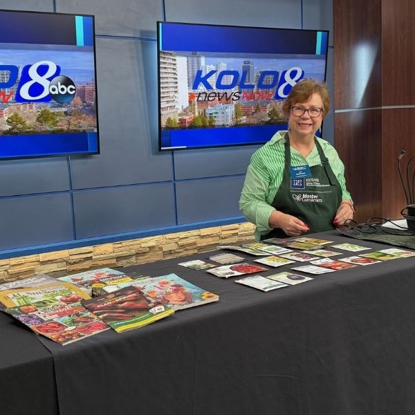 Master Gardener posing behind table with seed packets at news station KOLO