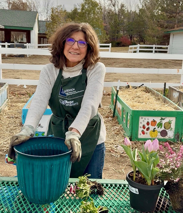 Person standing at a green picnic table holding a green pot and wearing a green apron with other potted flowers on the table. White stables in the background. Green trees in the background.