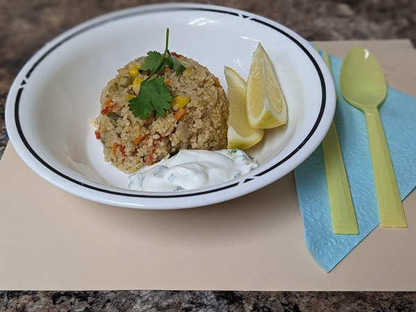 bowl of rice looking food with vegetables, lemmon slices, and yogurt next to it. Green garnish on top. Yellow silverware next to it.