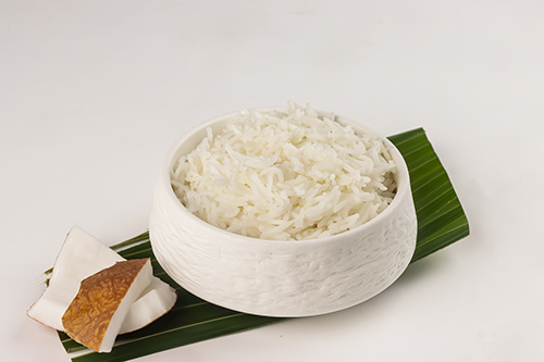 A white bowl with shredded white coconut on top of a green leaf and coconut pieces on the side.