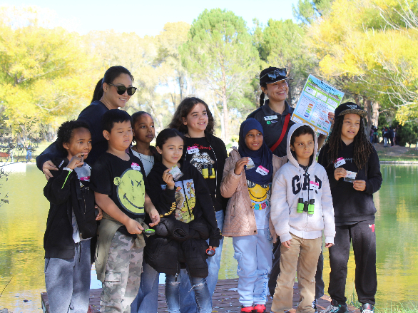 A group of youth and chaperones stand and pose for a picture on a dock with a pond in the background. The youth are holding up water samples they collected.