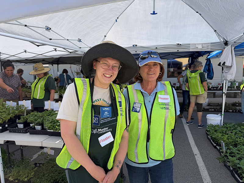 two people wearing neon colored vests under white tent surrounded by plants in trays