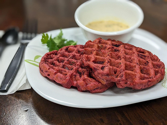red colored waffles on white plate with green garnish. yellow dipping sauce on side, black fork, spoon and napkin on side. 