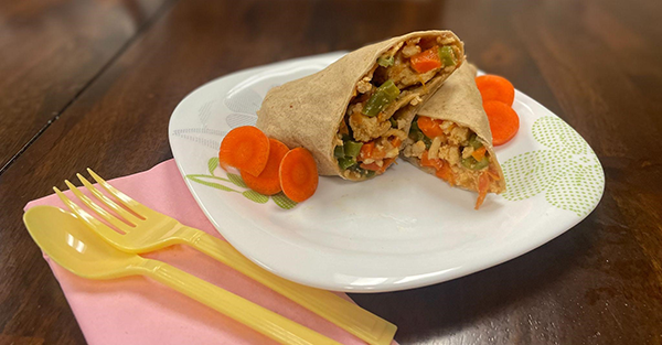 tortilla wrapped with vegetables in the middle and sliced carrots on the side on a white plate. Yellow silverware on the side.