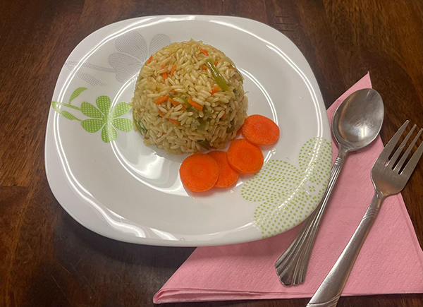 Scoop of rice with vegetables on a white plate and sliced carrots on the side. Silverware on the side.