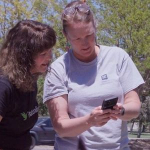 Two women look on a phone screen while standing in a garden