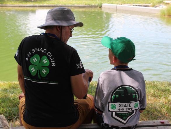 One adult and one youth sitting side by side by the pond at the 4-H Camp Alamo. Both individuals are wearing 4-H branded t-shirts.