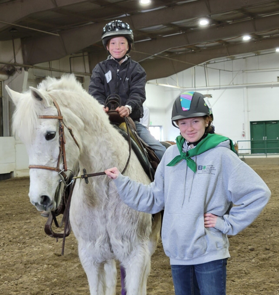 A young 4-H club member sits astride a white pony. They are wearing a dark jacket and a black riding helmet. They smile and hold the saddle. To the right stands an older 4-H club member who smiles with their hand on their hip. She is wearing a riding helmet, a green 4-H kerchief, and a grey sweater.