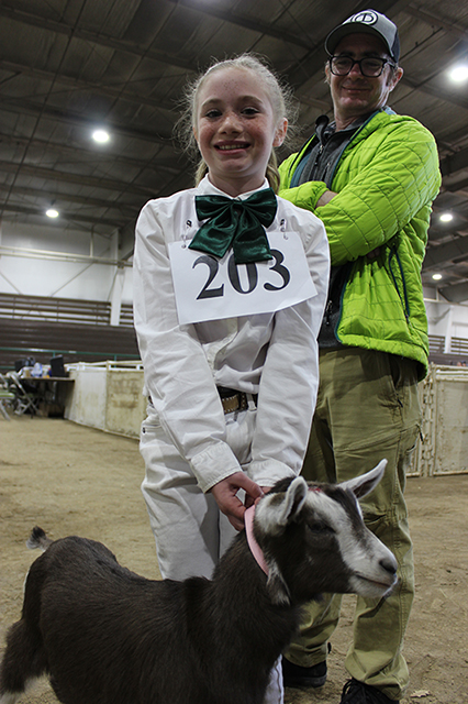 A youth in 4-H dress and sporting a contestant number holds her brown and white goat by its pink collar. Behind her, a man beams proudly.